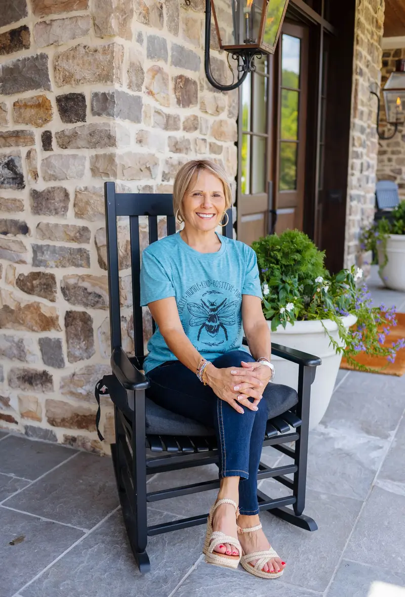 Heather Killingsworth in a Simply Busy Bee shirt, sitting in a rocking chair on a stone porch